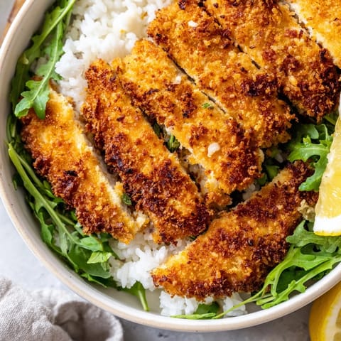 Slice of lemon resting on rice beside golden Parmesan chicken cutlet bowl topped with arugula.