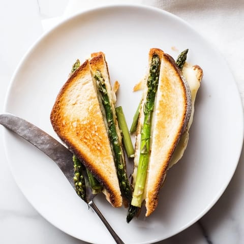A close-up of the Asparagus & Brie Grilled Cheese reveals gooey, creamy cheese oozing from buttery, golden-toasted bread layered with tender green asparagus.  