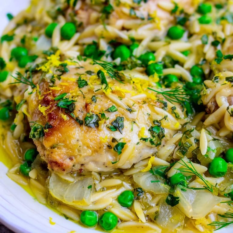 This Mediterranean-inspired Lemon Herb Chicken Orzo Skillet features juicy chicken thighs, al dente pasta, and vibrant peas, garnished with herbs and a lemon wedge on a rustic wooden table.