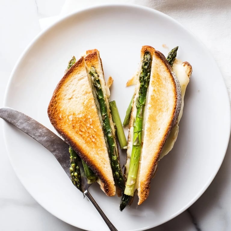 A close-up of the Asparagus & Brie Grilled Cheese reveals gooey, creamy cheese oozing from buttery, golden-toasted bread layered with tender green asparagus.  